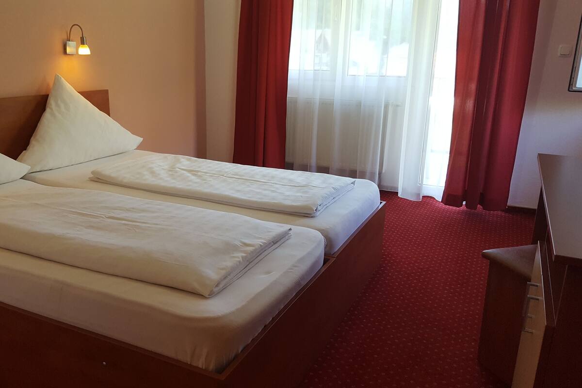 Ein Hotelzimmer mit zwei Betten, einem Holzkopfteil, einem roten Teppich und einem Fenster mit roten Vorhängen.