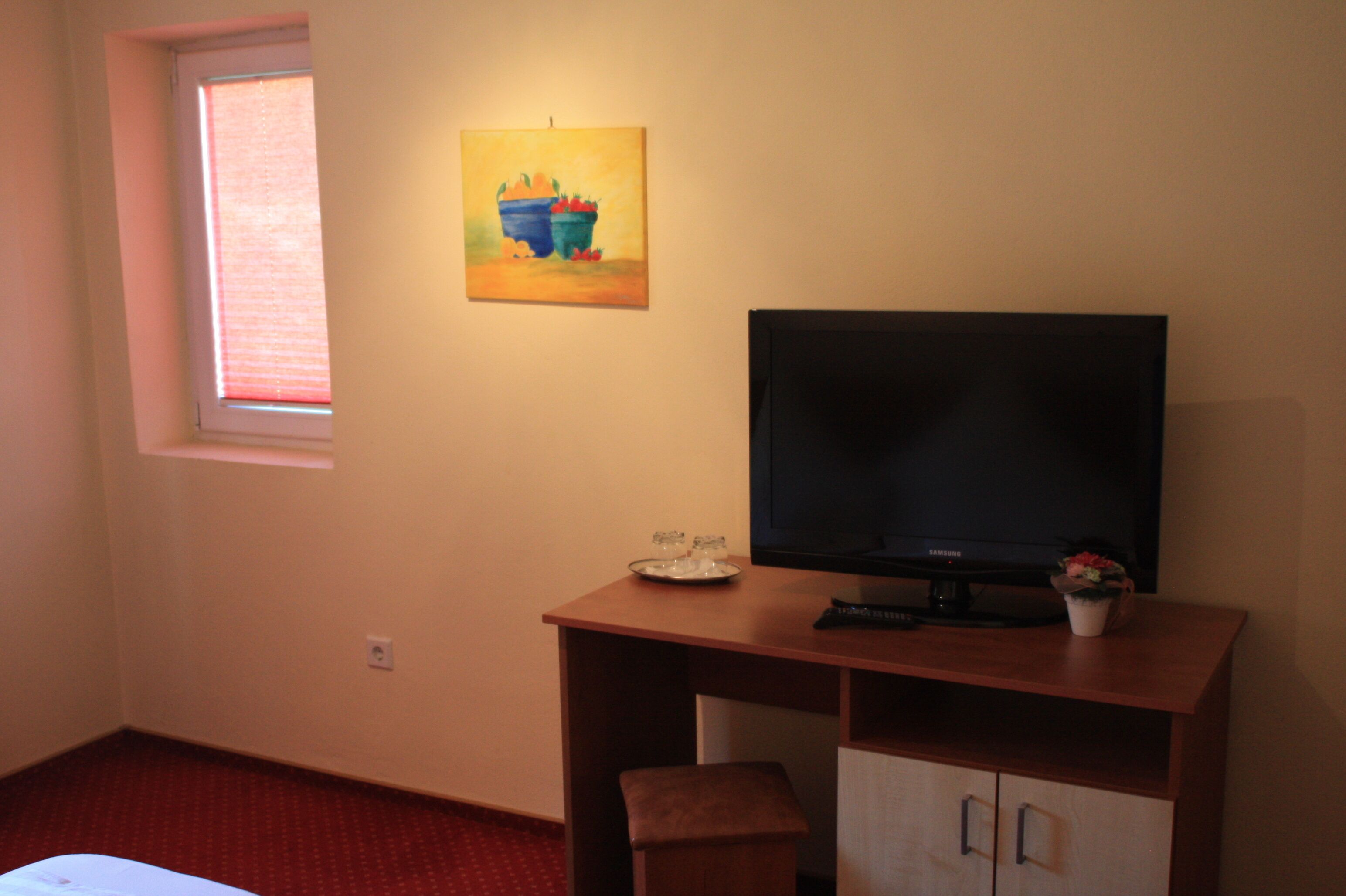 Una habitación pequeña con una televisión sobre un escritorio de madera, una ventana con persianas y un cuadro en la pared.