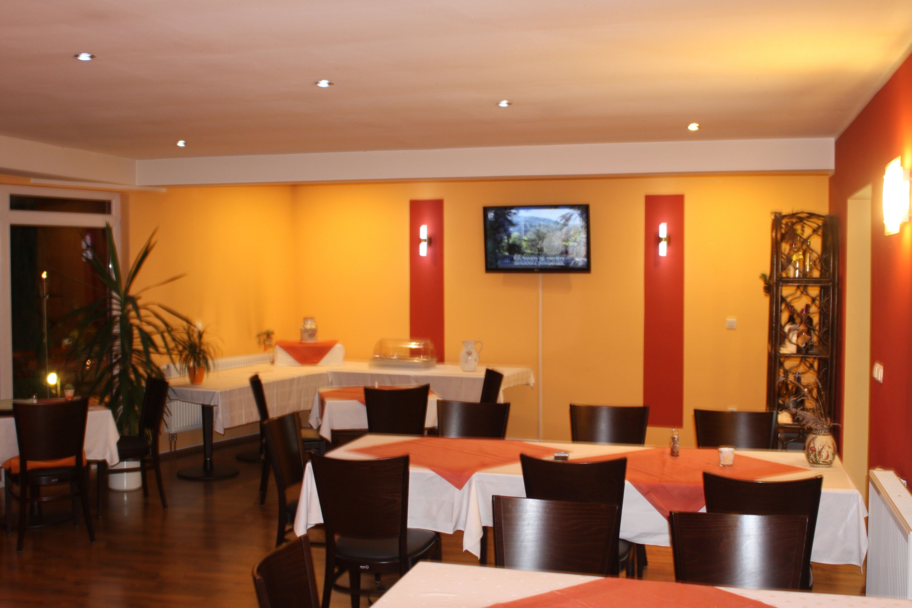 A restaurant with tables set for dining, a television mounted on the wall, and a plant in the corner.