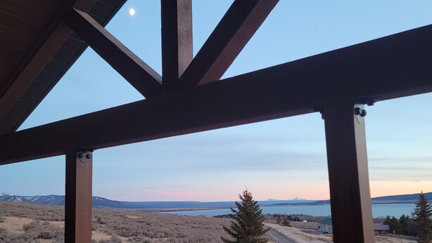 A view through a wooden-framed window overlooking a landscape with a body of water, distant mountains, and sparse vegetation.