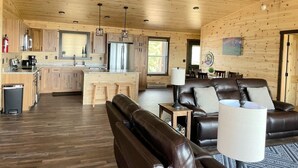 A spacious living area with a brown leather sofa, a coffee table, and a dining table with chairs. There is a kitchen with wooden cabinets and a refrigerator.