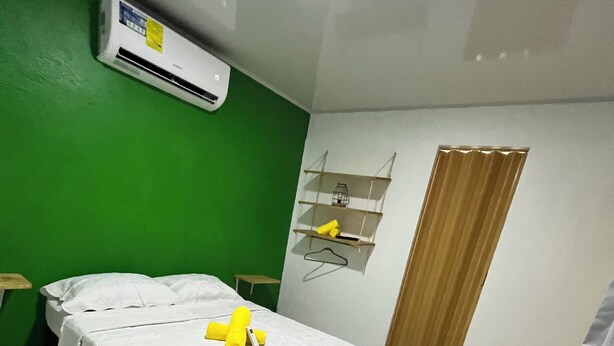 Una habitación con una cama, una silla, una mesita, un estante con objetos y una puerta.