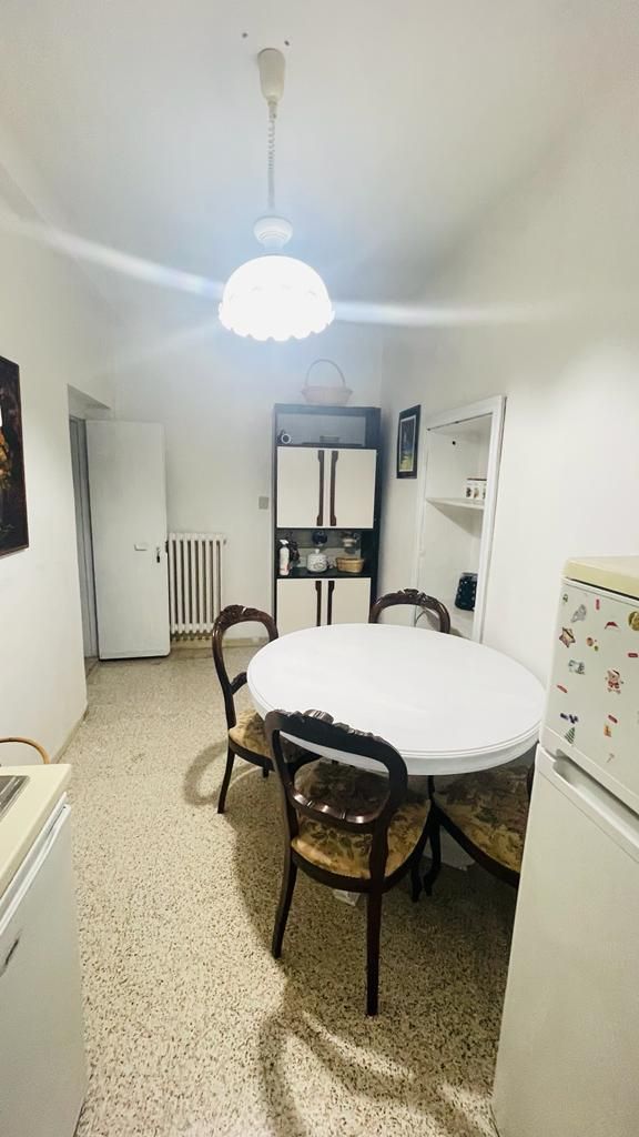 A compact dining area with a round table, four chairs, a ceiling fan, a radiator, and a refrigerator.