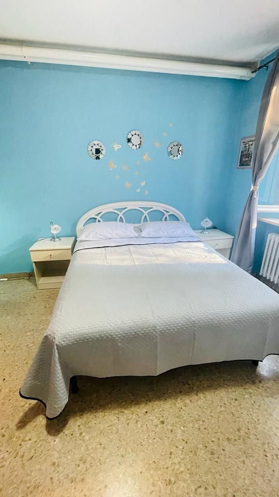 A bedroom with a bed, two bedside tables, a wall clock, and a window with curtains.