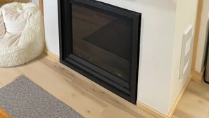 A modern living room with a fireplace, a white fluffy chair, and a small shelf with decorative items. - Lake Front Cottage on Trooper Lake, Troy Hill Ont (Tory Hill)