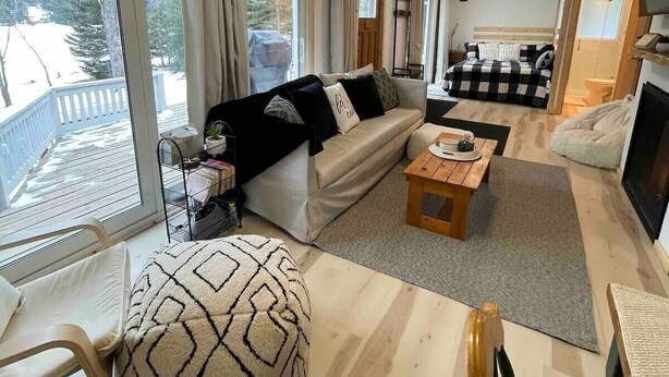 A cozy living room with a sofa, a coffee table, and a patterned ottoman. There is a dining area with a table and chairs, and a large window offering a view of a snowy deck. - Lake Front Cottage on Trooper Lake, Troy Hill Ont (Tory Hill)