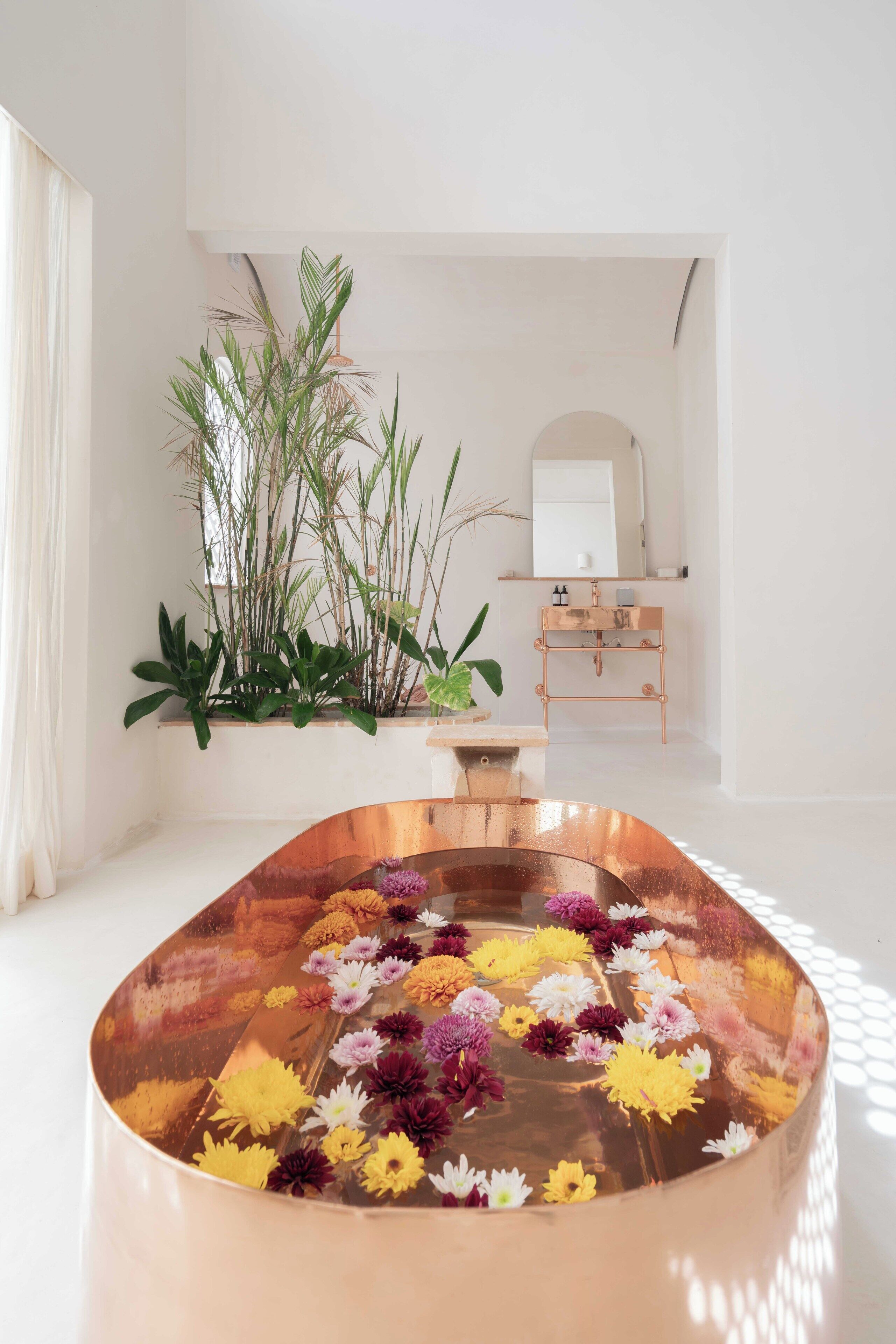 Un baño moderno con una bañera de cobre llena de flores, un lavamanos blanco y un espejo encima.
