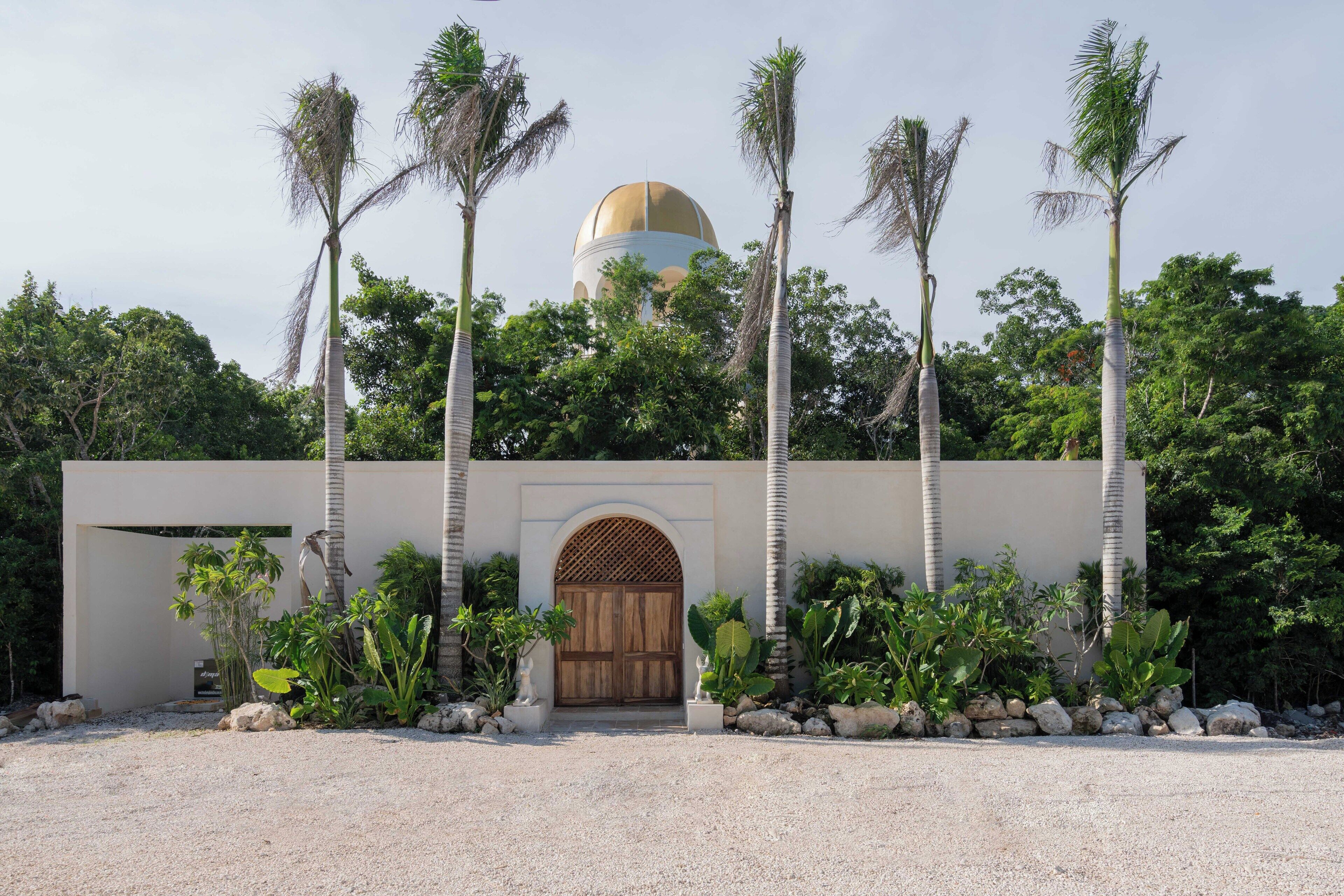 A building with a large golden dome, surrounded by palm trees and a gravel pathway leading to a wooden door.