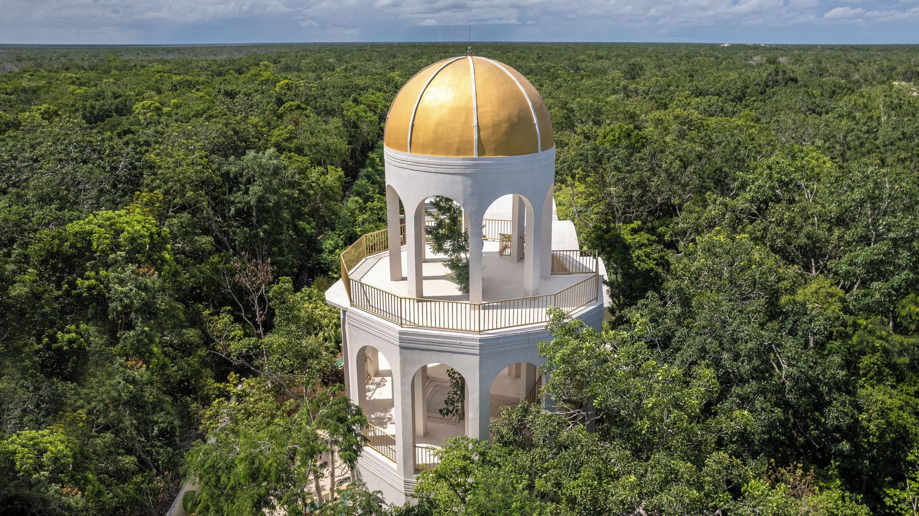 A white tower with a golden dome, surrounded by dense green forest.