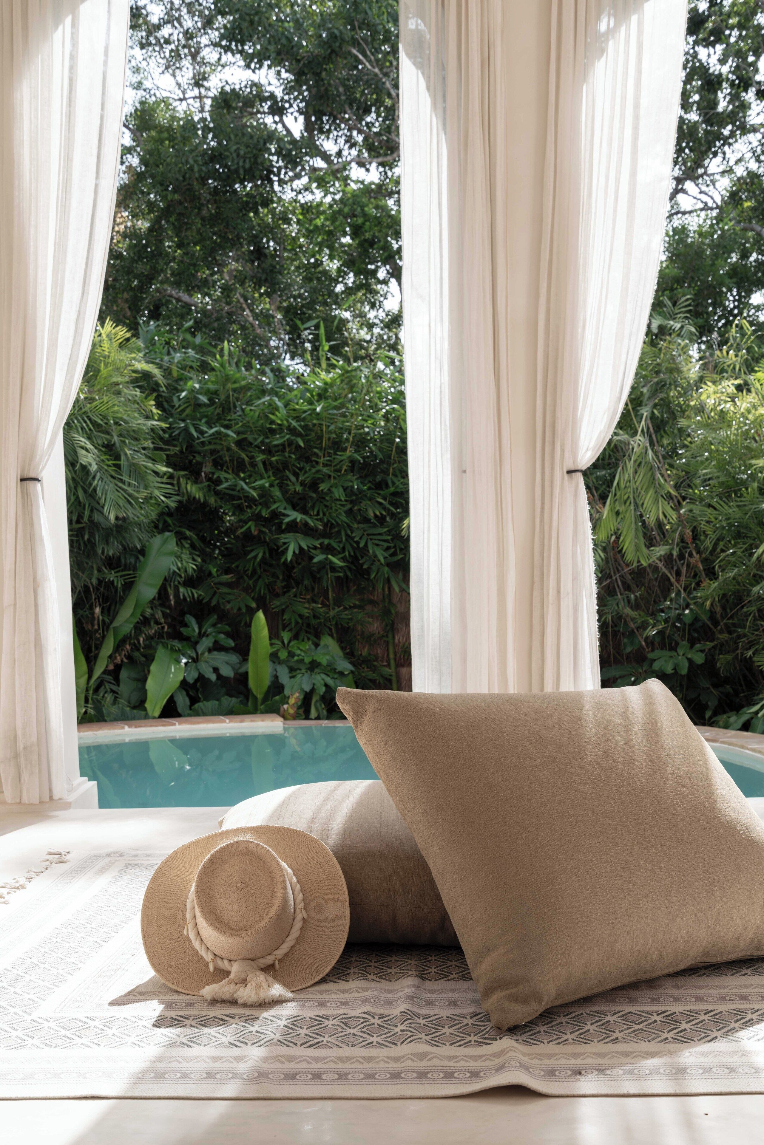 Una cama con un almohadón color crema y un sombrero de paja, frente a un fondo de vegetación y una piscina.