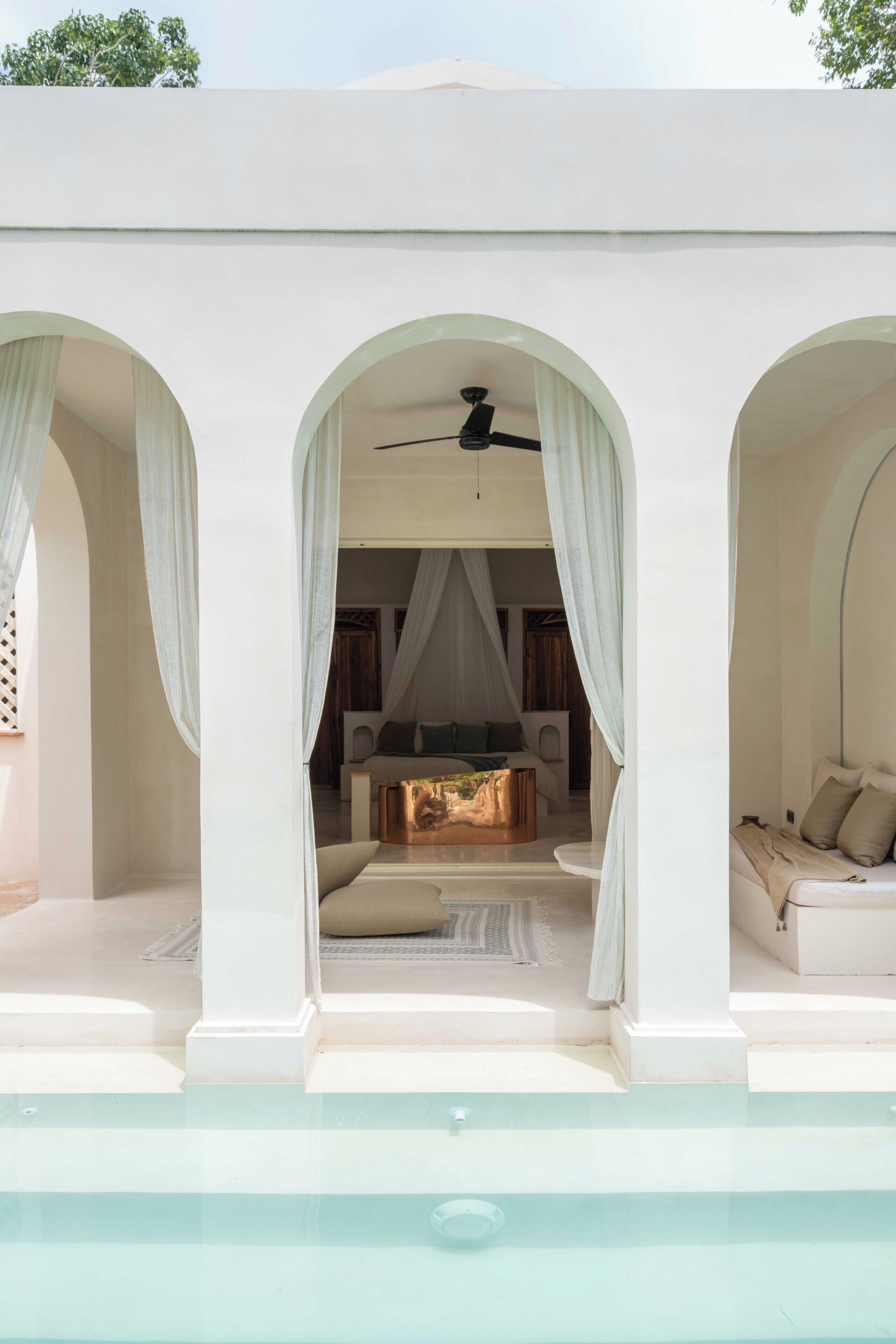 Un espace piscine doté d’un coin salon aménagé sous des arcades, d’un ventilateur de plafond et d’une vue sur l’intérieur.