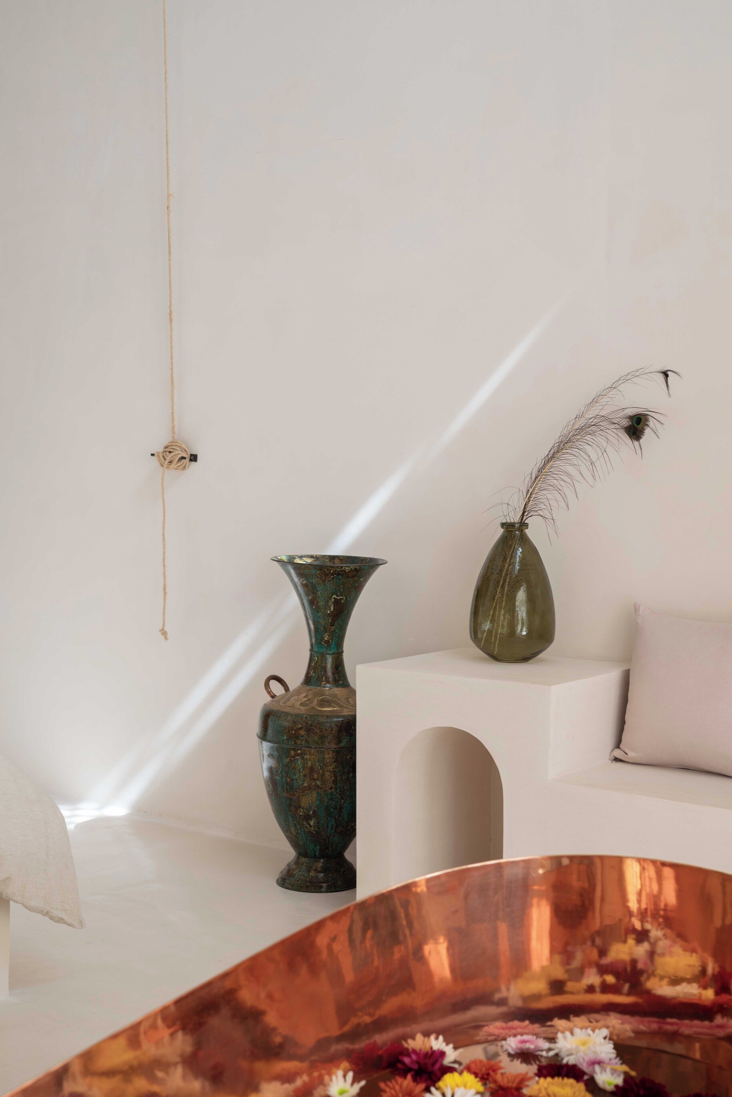 A minimalist room with a white sofa, a green vase with a feather, and a copper bowl with flowers.