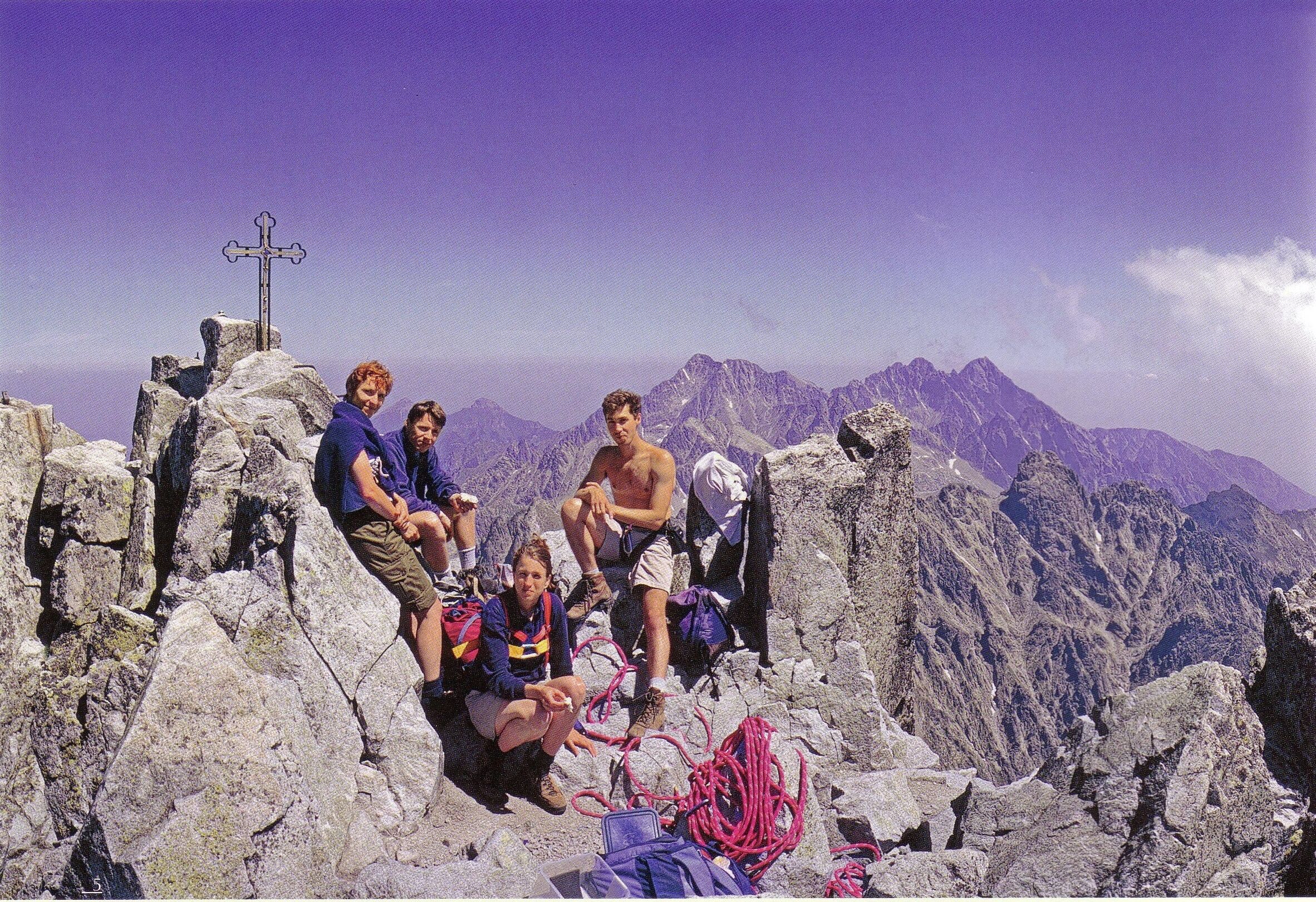 Un groupe de personnes se trouve au sommet d’une montagne, dans un paysage rocheux, près d’un monument en forme de croix.