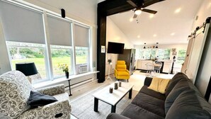 A modern living room with a sofa, armchair, and a yellow chair. There is a kitchen in the background with white cabinets and a dining area. - The Barnhouse - Country Charm 2 miles from Colgate, renovated century-old barn (Hamilton)