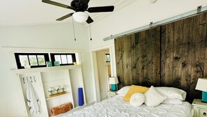 A bedroom with a bed, a ceiling fan, a wooden headboard, a nightstand, a lamp, a bench, and a shelf with decorative items. - The Barnhouse - Country Charm 2 miles from Colgate, renovated century-old barn (Hamilton)