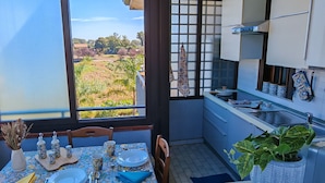 A dining area with a table set for two, a kitchen with white cabinets and blue tiles, and a view of a green landscape through a window. - Blue Garden Home - Fondachello (Mascali)