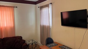 A living room with a sofa, a glass-top coffee table, a TV mounted on the wall, and a window with curtains. - Joy Apartment #3 centrally located.  (Anniston)