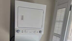 A stacked washer and dryer unit in a room with a wooden floor and a white door with blinds. - Joy Apartment #3 centrally located.  (Anniston)