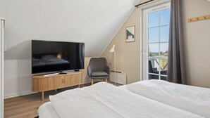A modern hotel room with a TV, a wooden side table, a grey armchair, a large bed, and a view of the outdoors through a window. - Cozy, central house near the sea (Brenderup)