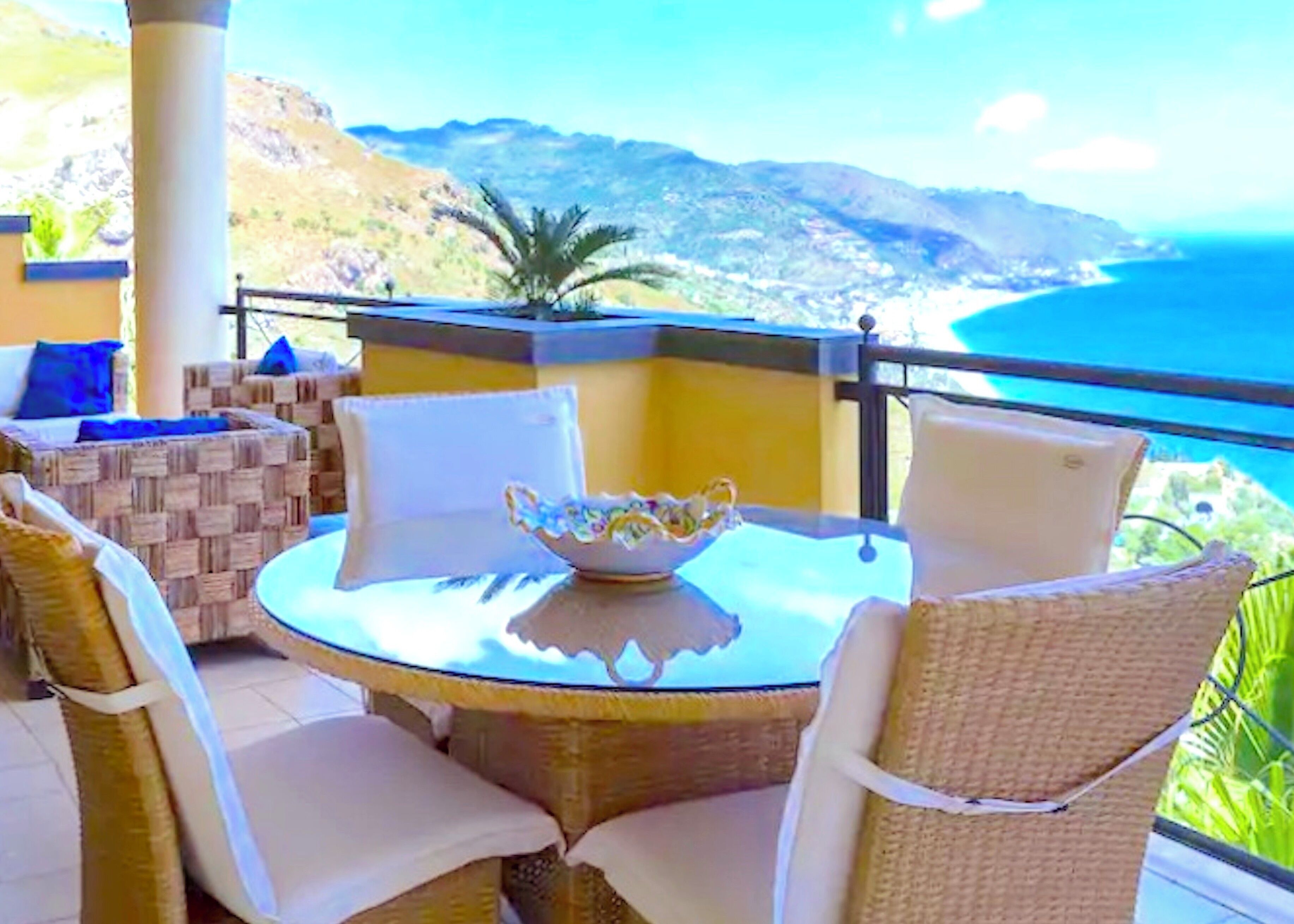 Ein runder Tisch mit einer Schüssel, umgeben von Korbstühlen mit weißen Kissen, auf einem Balkon mit Blick auf das Meer und die Berge.