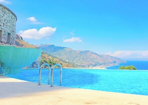 Ein Pool mit klarem, blauem Wasser, einer Steinsäule und Blick auf eine gebirgige Küstenlinie.