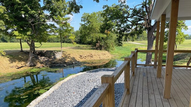 A wooden deck with a stone border overlooking a calm pond and a grassy field.