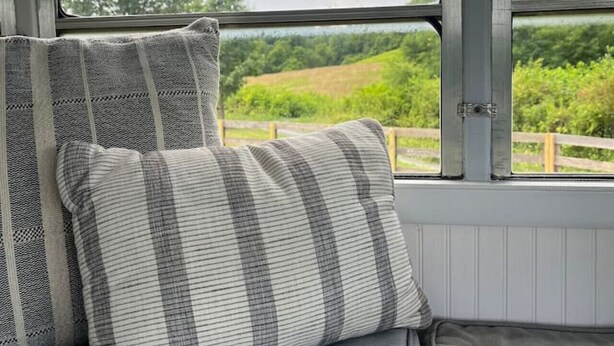 A bed with grey bedding and two pillows, a window with a view of greenery, and a metal structure above the window.
