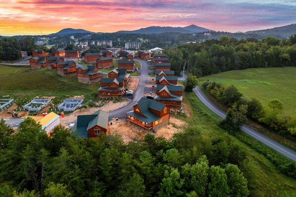 A cluster of log cabins with a dirt road leading to a main entrance, surrounded by dense forest and a mountain in the background.