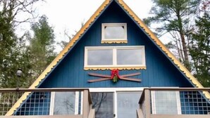 A two-story house with a blue exterior, a wooden staircase leading to the upper floor, and a stack of firewood on the ground floor. - Direct Waterfront, Year-Round, A-Frame Cabin (Antrim)