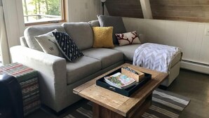 A cozy living room with a grey sectional sofa, a wooden coffee table with magazines, and a view of the outdoors through a large window. - Direct Waterfront, Year-Round, A-Frame Cabin (Antrim)