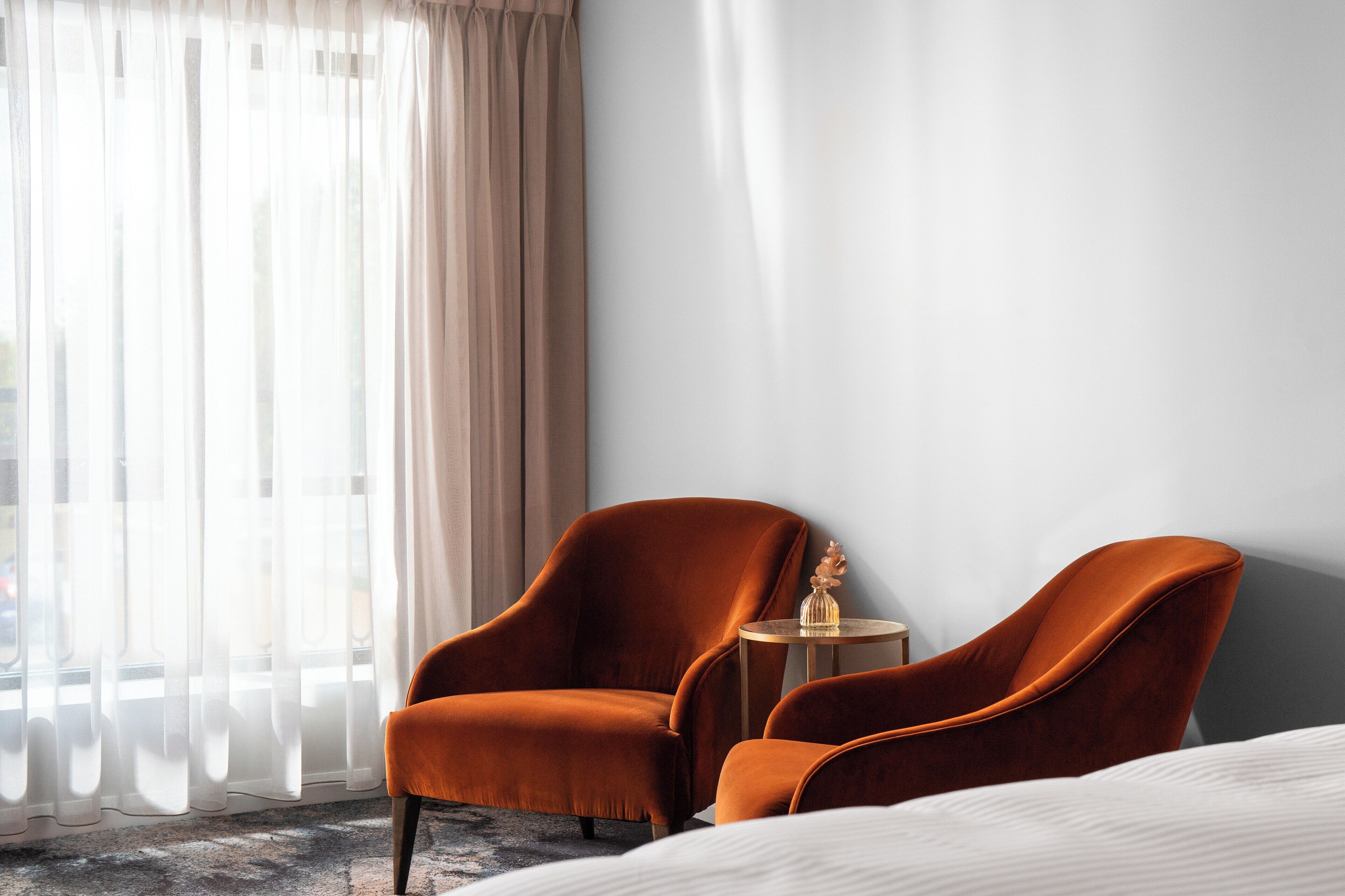 A hotel room with a bed, two orange armchairs, a small round table, and a large window with sheer curtains.