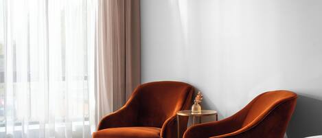 A hotel room with a bed, two orange armchairs, a small round table, and a large window with sheer curtains.