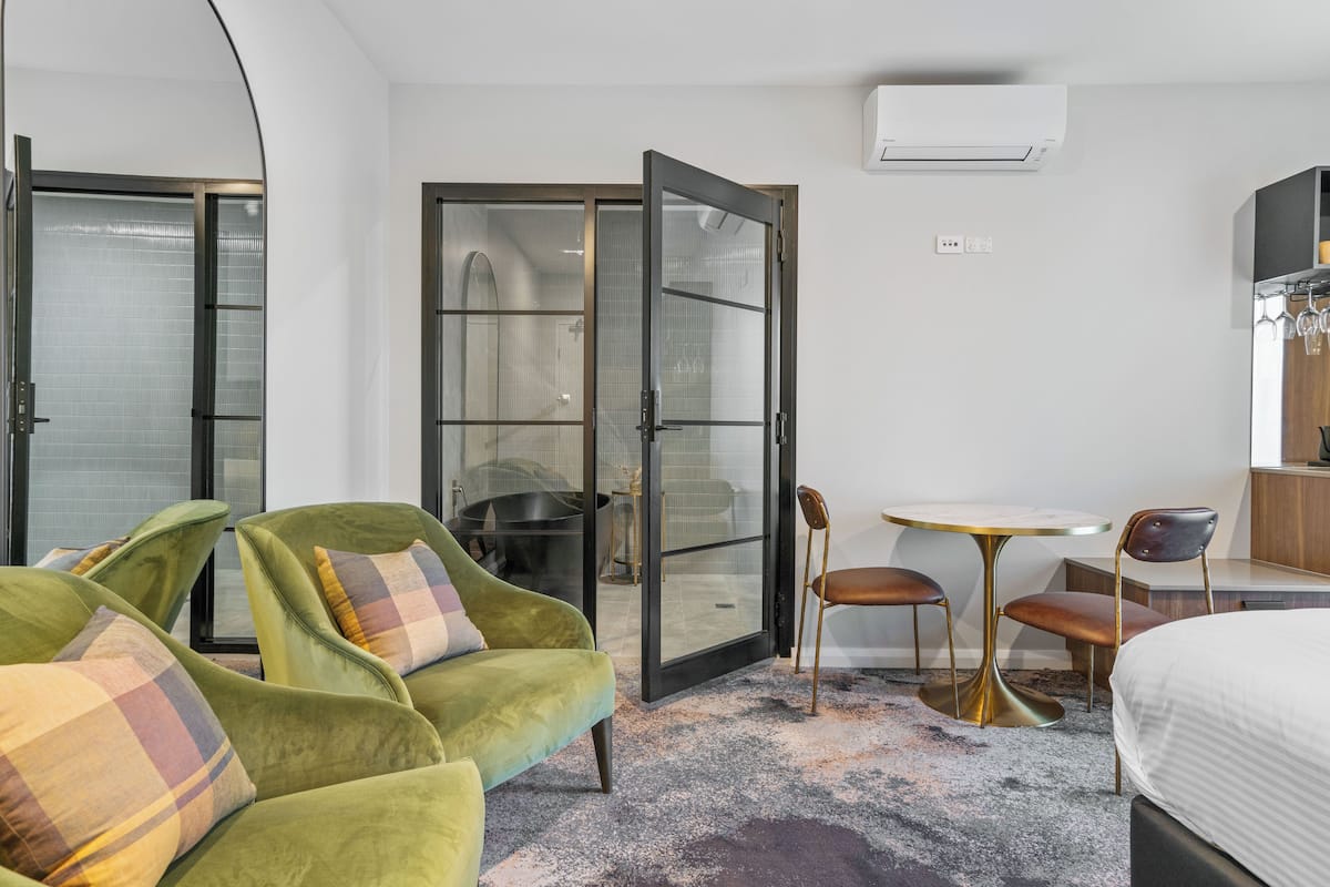 A hotel room with a bed, a green armchair with a plaid cushion, a small round table with two chairs, and a sliding glass door leading to a bathroom.