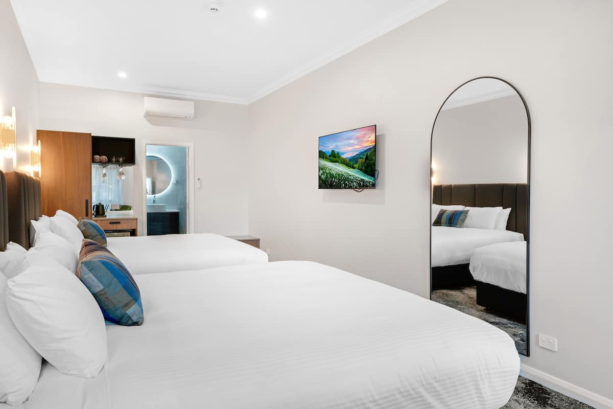 A hotel room with two beds, a large mirror, a painting, and a modern bathroom with a circular mirror.