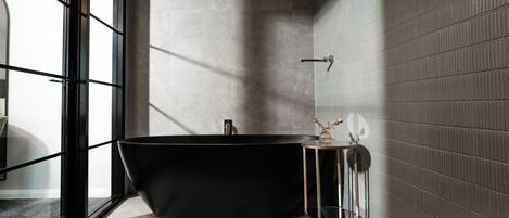 A modern bathroom with a freestanding bathtub, a small table with a vase, and a window providing natural light.