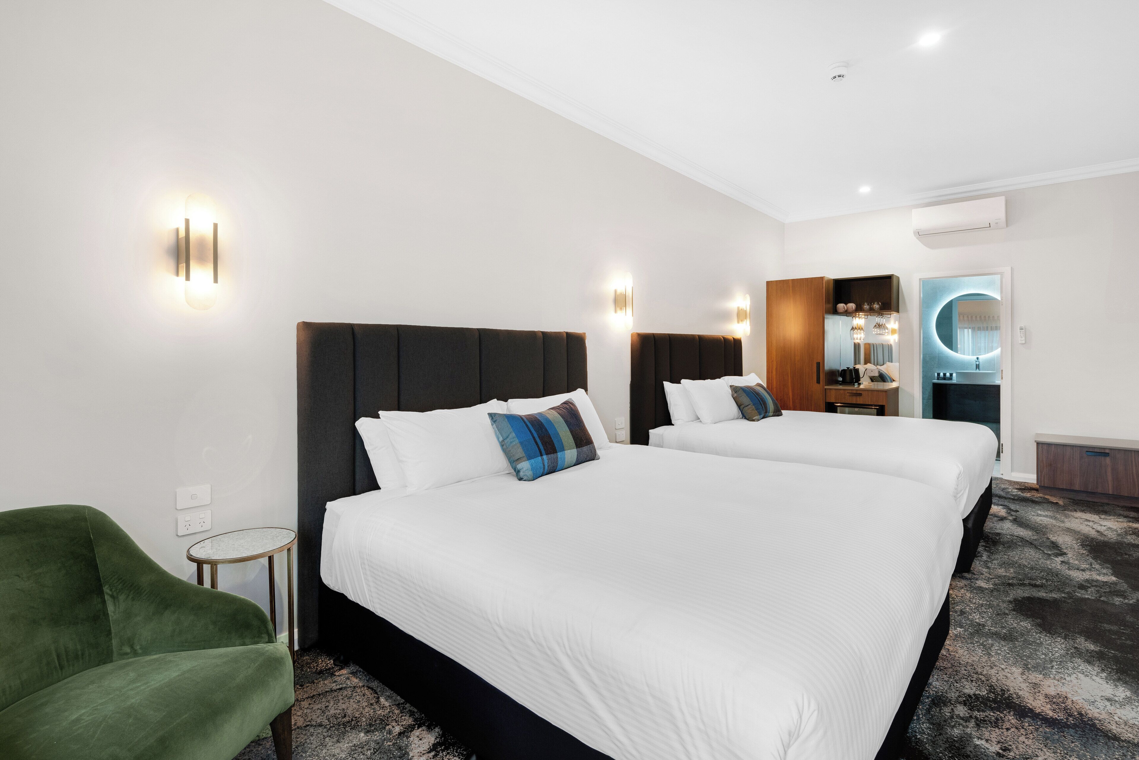 A hotel room with two beds, a green armchair, a small side table, and a bathroom visible through an open door.