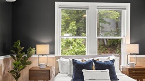 A bedroom with a large window, a bed with white and blue bedding, two bedside tables with lamps, and a potted plant.