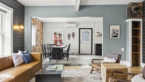 A modern living room with a sofa, coffee table, and a fireplace. There is a dining area with a table and chairs. The room has a neutral color palette and features a brick accent wall.