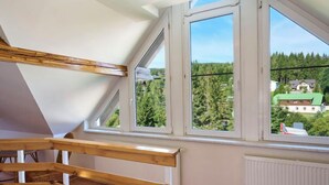 A room with a sloped ceiling, wooden staircase, and large windows offering a view of a forest and buildings. - Harrachov 497 by Interhome (Harrachov)