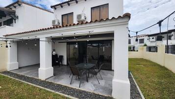 A two-story house with a patio area featuring a table and chairs, air conditioning units, and a pergola with string lights.