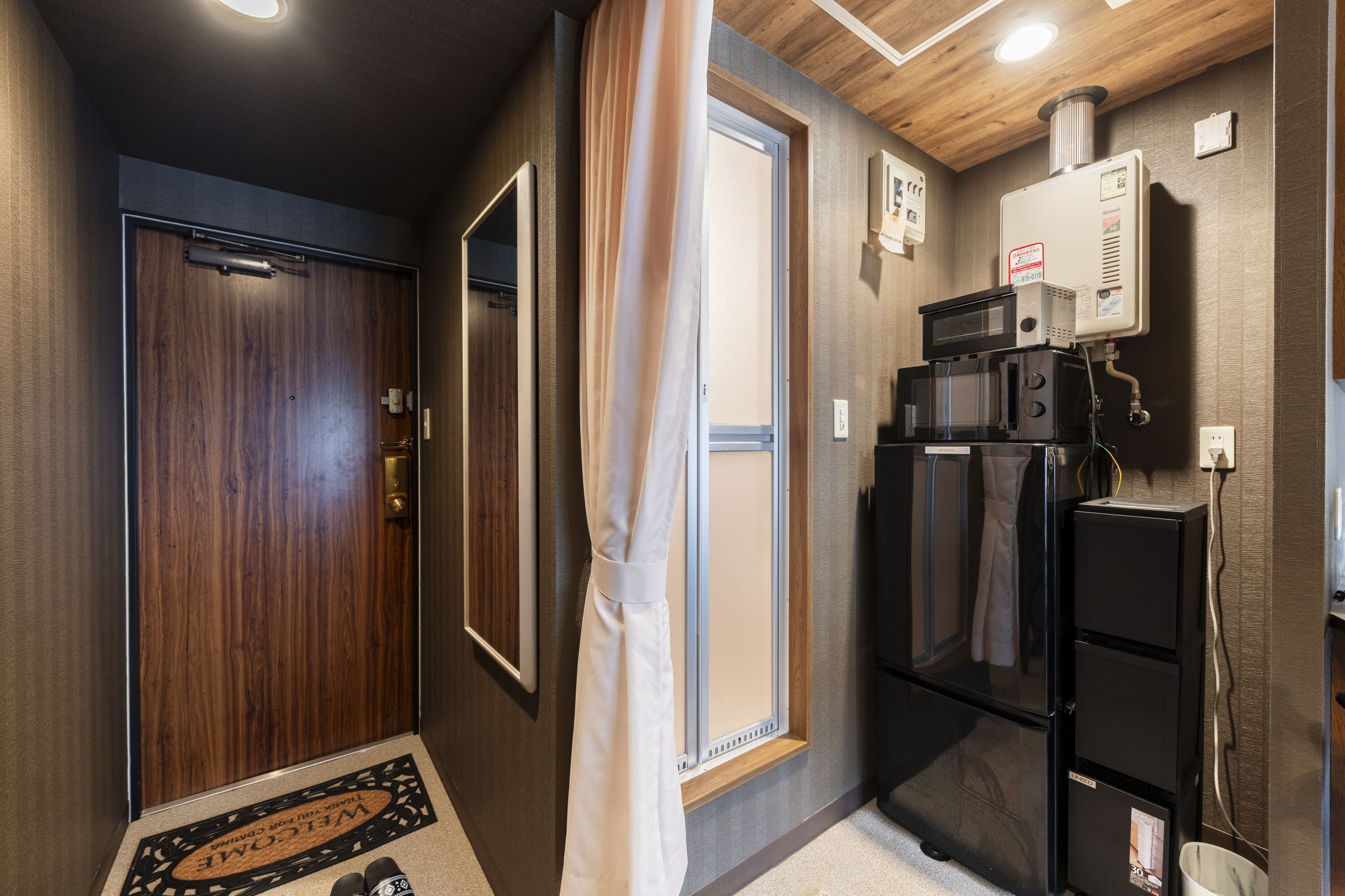 A compact hotel room with a wooden door, a sliding glass door, a black refrigerator, a microwave, and a water heater on the wall.