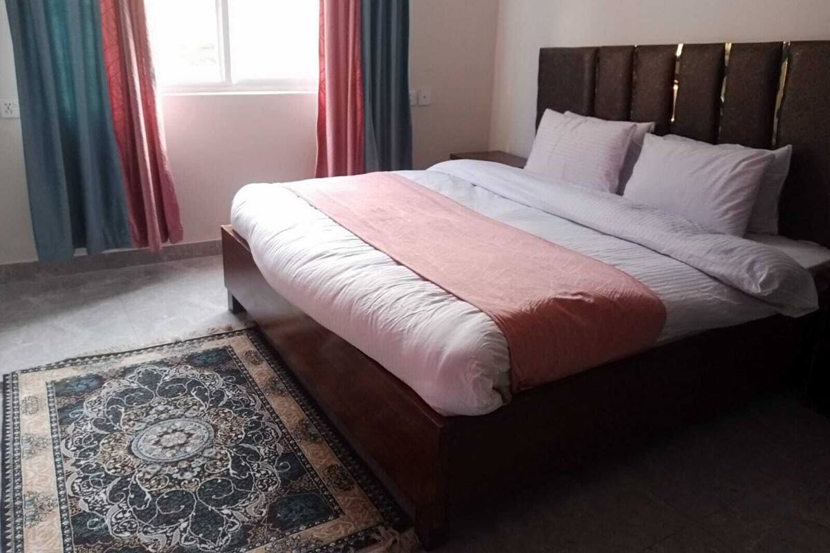 A neatly made bed with a headboard, a patterned rug, and curtains on a window.