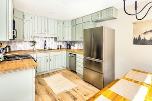 A kitchen with light green cabinets, a stainless steel refrigerator, wooden countertops, and a wooden floor.