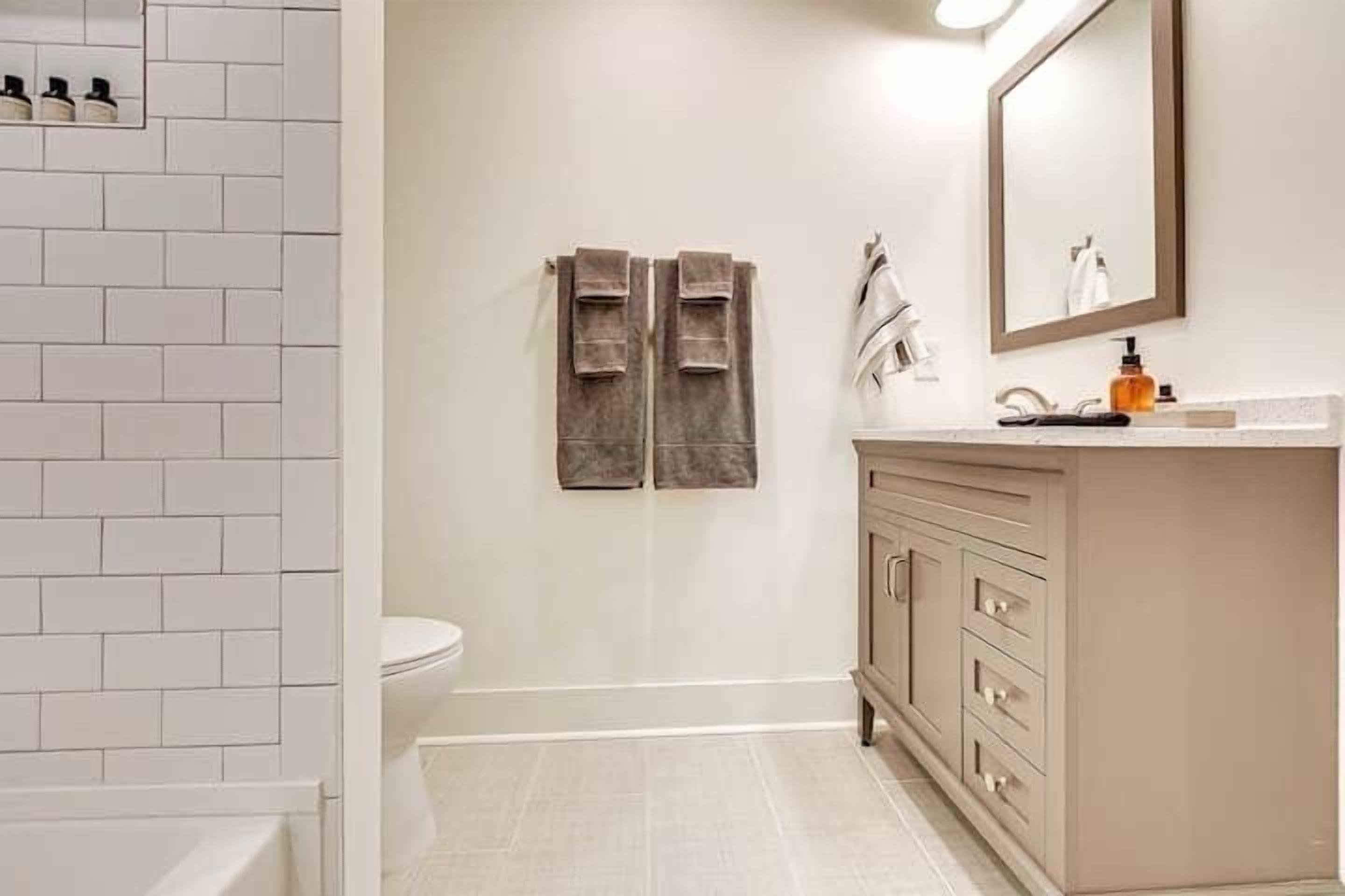 Une salle de bain aux murs recouverts de carreaux blancs, un meuble-lavabo beige avec des tiroirs, un miroir et deux serviettes suspendues à un porte-serviettes.