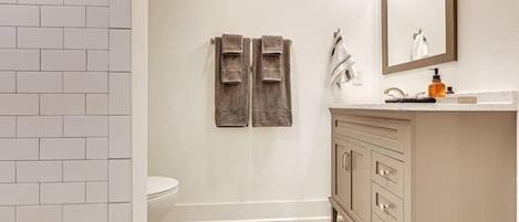 Une salle de bain aux murs recouverts de carreaux blancs, un meuble-lavabo beige avec des tiroirs, un miroir et deux serviettes suspendues à un porte-serviettes.