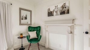 Une pièce avec un fauteuil vert, une petite table sur laquelle est posée une plante, une cheminée et deux tableaux encadrés accrochés au mur.