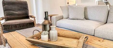 Una sala de estar con un sillón de cuero marrón, un sofá gris y una mesa de centro de madera con una bandeja que sostiene un florero y una planta suculenta.