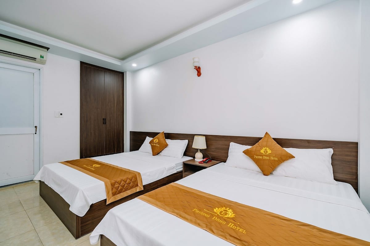 Quarto de hotel com duas camas, cada uma com colcha marrom e travesseiro com o logotipo do hotel. Entre as camas, há uma mesinha de cabeceira com abajur e telefone.