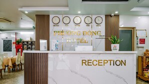Un espace d’accueil doté d’un comptoir en marbre, de plusieurs horloges indiquant l’heure à New York, Tokyo, au Vietnam, à Paris et à Londres, et d’une enseigne indiquant « RÉCEPTION DE L’HÔTEL PHUONG DONG ».