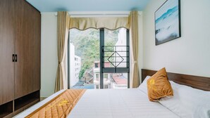 A hotel room with a bed, a wooden wardrobe, a view of buildings through a window, and a painting on the wall. - Phuong Dong Hotel (Hai Phong)